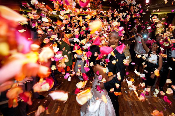 fun photo of  the bride and groom caught in storm of dark pink, orange, and yellow rose petals being thrown at them by family and guests during their first dance at the reception - photo by Houston based wedding photographer Adam Nyholt