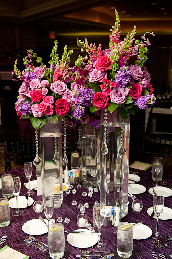 table setting at reception - three tall glass vases with danging crystals with dark pink, light pink, purple, and green floral arrangements as centerpiece and purple tablecloth - photo by Houston based wedding photographer Adam Nyholt