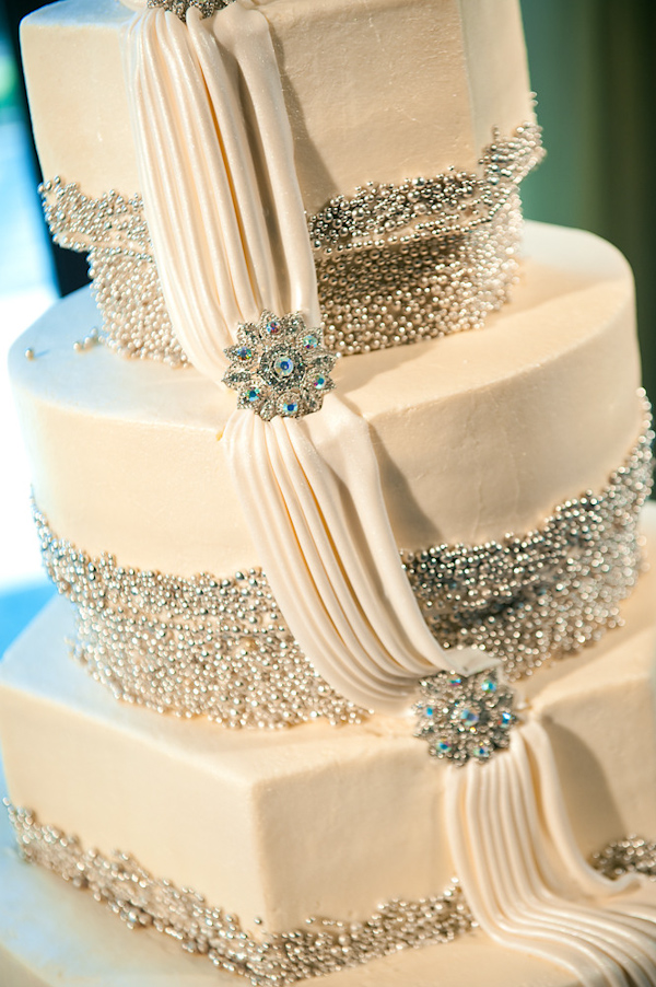beautiful ivory three tiered cake with silver beaded borders and a drape across all borders with silver and aqua accented stone brooches as cake topper - photo by Houston based wedding photographer Adam Nyholt