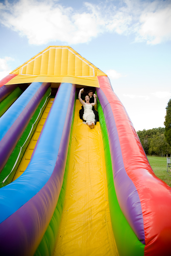 fun photo of bride and groom going down a colorful blow up slide with a blue sky and white clouds in the background - photo by North Carolina based wedding photographers Cunningham Photo Artists