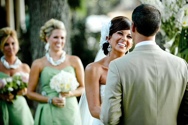 photo of the beautiful bride smiling at the groom during the ceremony with the bridesmaids behind her - bride is wearing white hairpiece with full length veil and chandelier earrings and bridesmaids are wearing green dresses -  photo by North Carolina based wedding photographers Cunningham Photo Artists