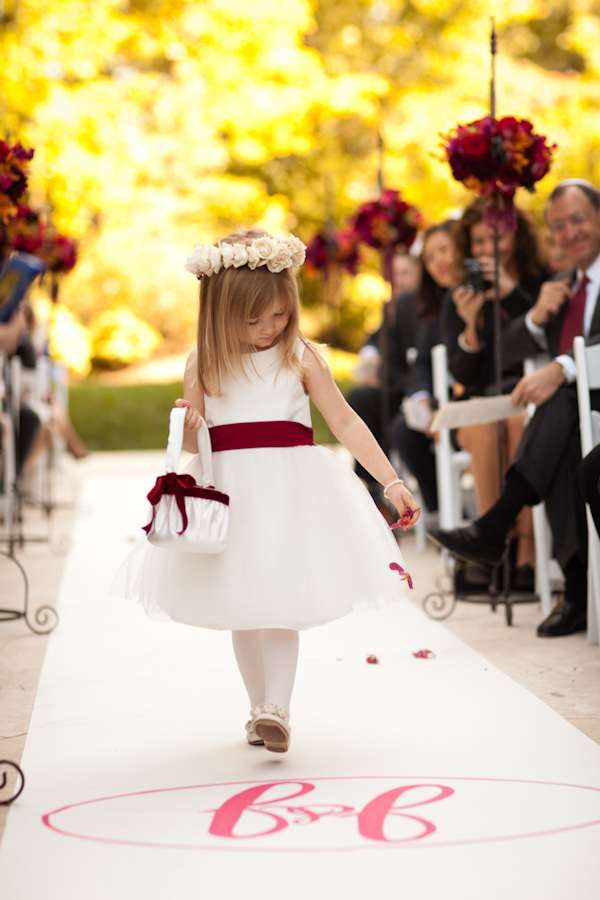 adorable flower girl wearing a white dress with a dark red sash carrying a white basket with dark red decor down the ceremony aisle - photo by Washington DC based wedding photojournalist Paul Morse