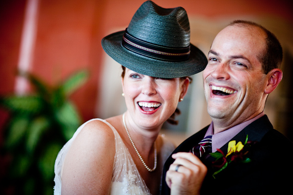 the happy couple laughing together as the bride is wearing the groom's black hat with black and purple border - bride is wearing pearls and a white dress and the groom is wearing a black suit with a purple shirt and yellow and dark pink boutonniere - photo by New Mexico based wedding photographers Twin Lens