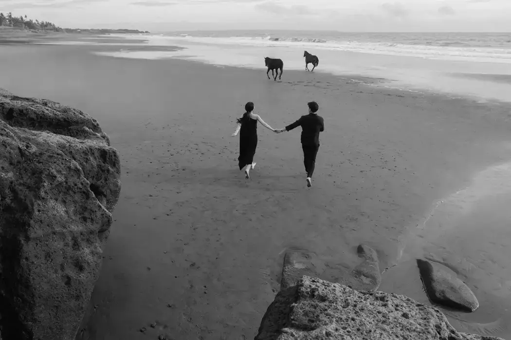 black and white photo couple running on beach holding hands with two wild horses