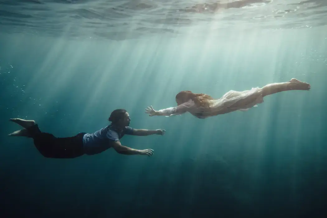 man and woman underwater swimming toward each other with sunlight coming through