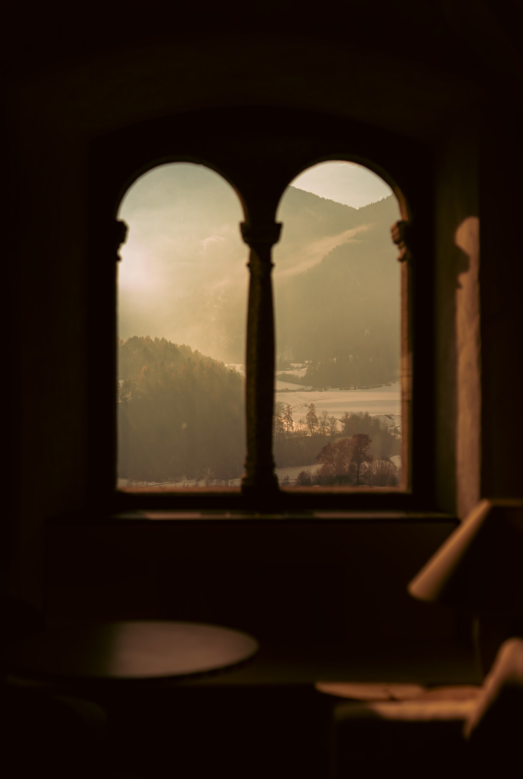 Arched double window in a dim castle room at Castel Badia, framing a hazy golden view of forested mountains and the snowy valley below