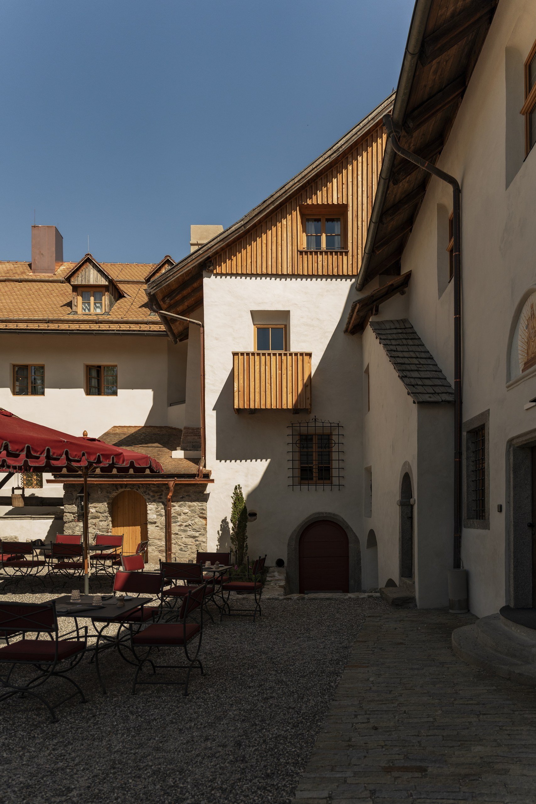 Castle courtyard at Castel Badia with white stone walls, a wooden balcony, and terrace tables under red umbrellas on a sunny spring day