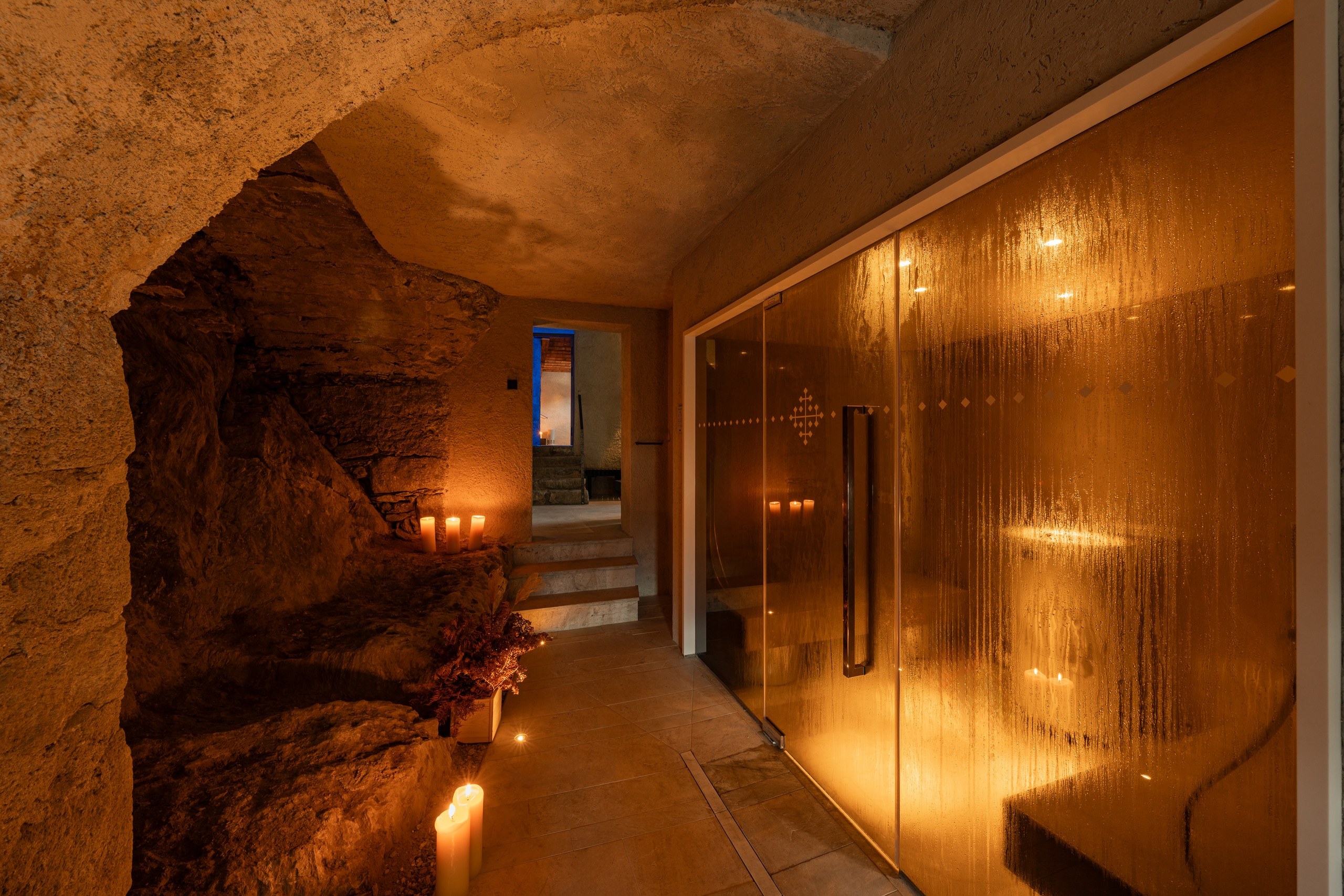 Candlelit spa corridor at Castel Badia with rough rock walls on one side and a glass-walled sauna glowing amber on the other