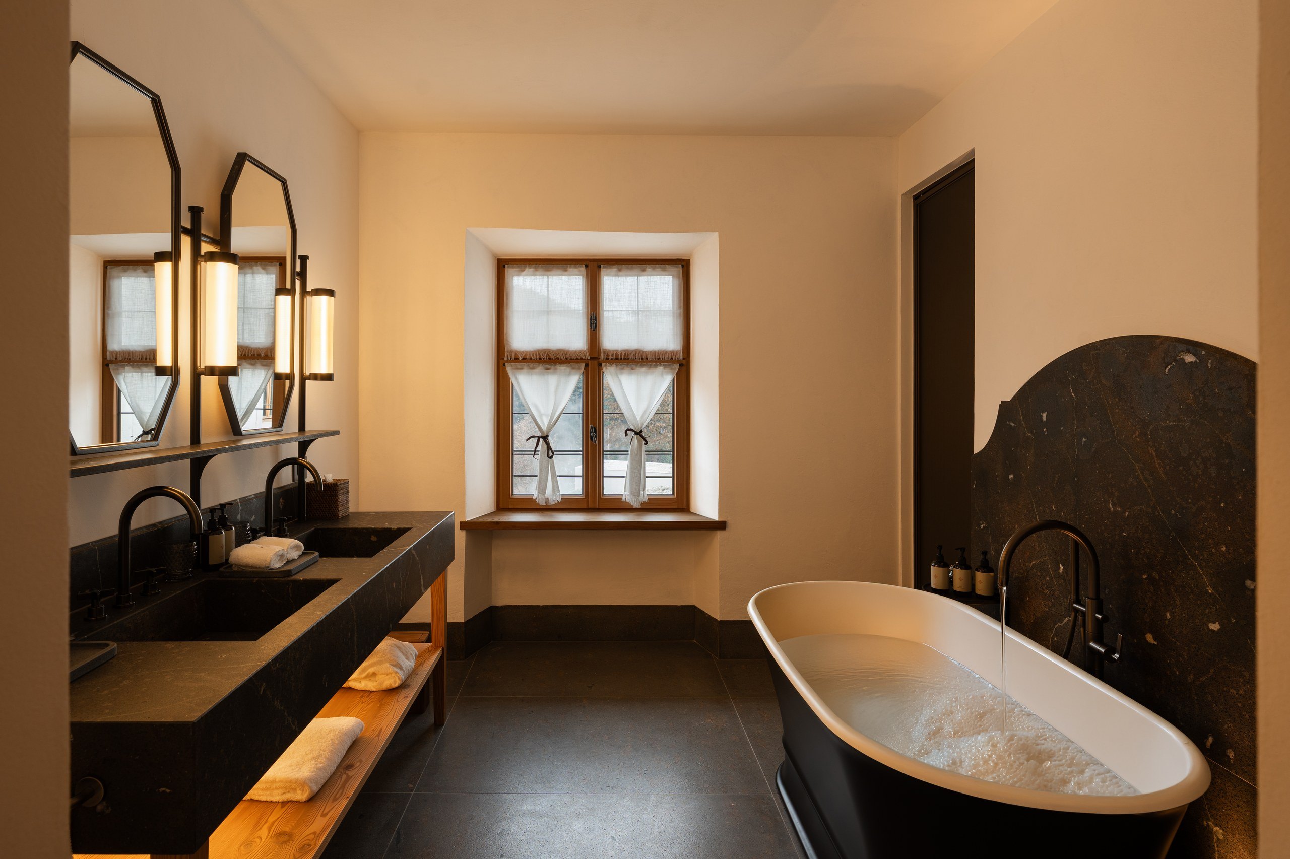 Suite bathroom at Castel Badia with a freestanding soaking tub, dark stone double vanity, and a window with mountain views