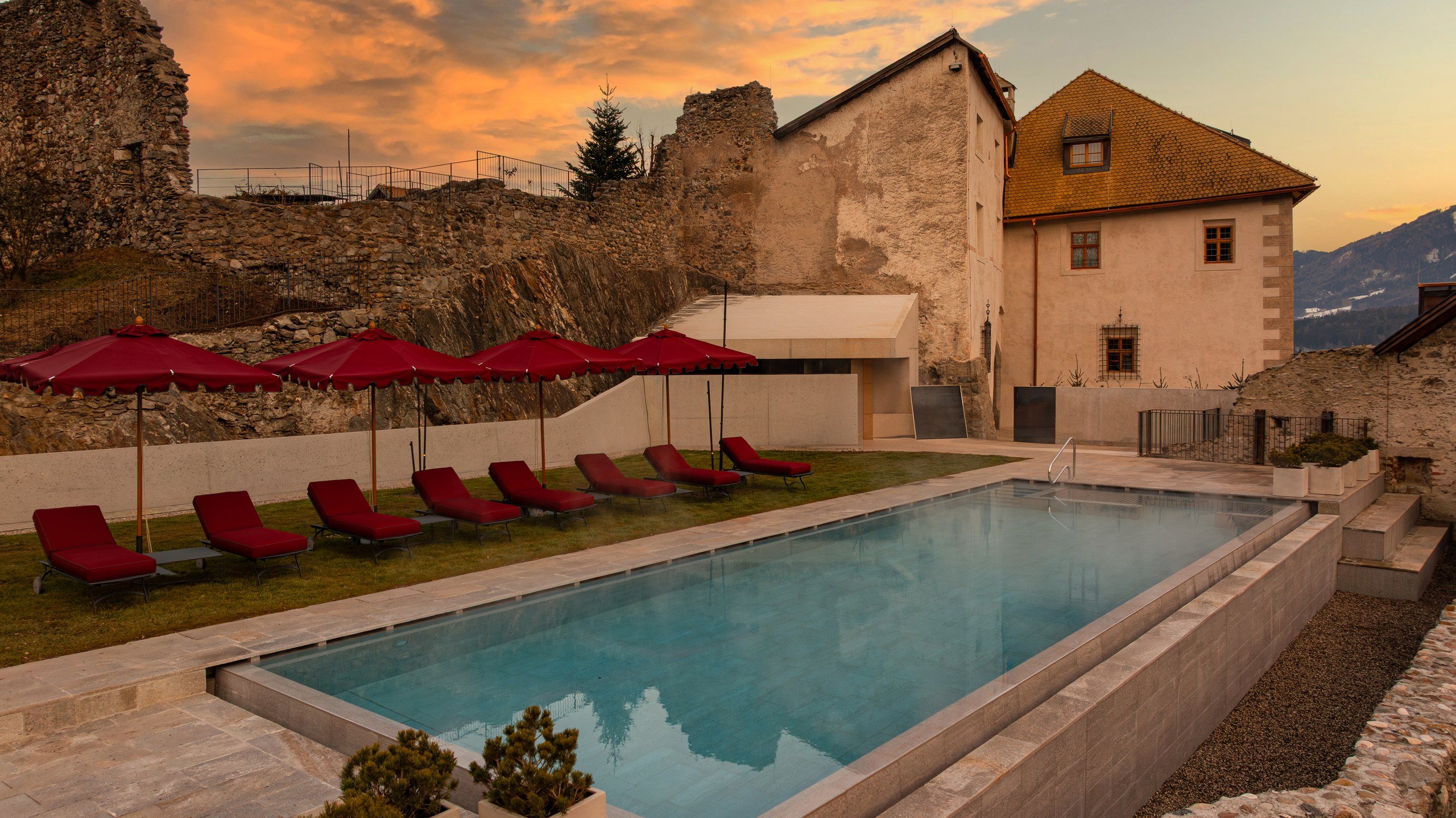 Outdoor pool with red loungers and umbrellas beside castle ruins at sunset at a Dolomites castle hotel