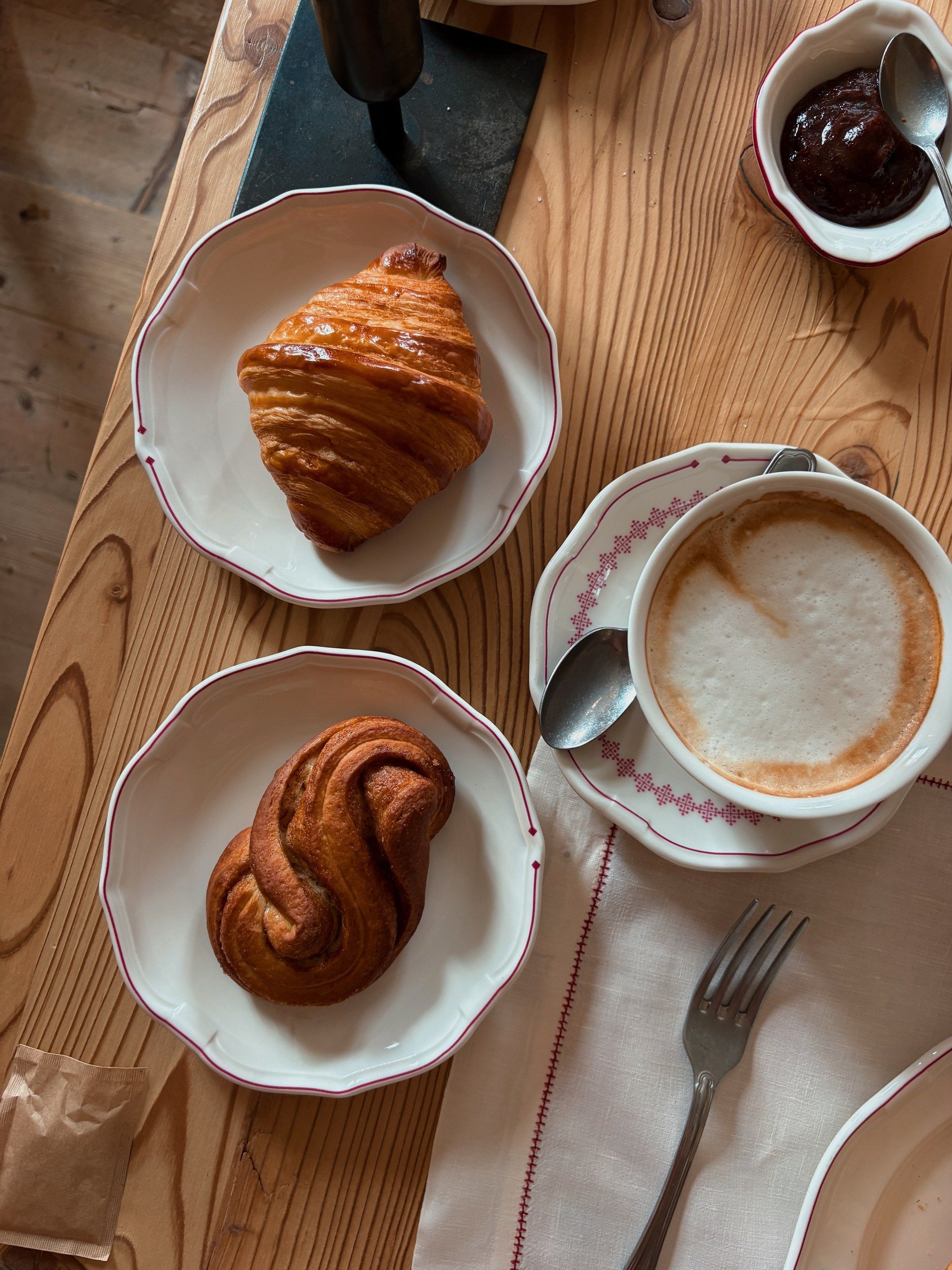 Hotel breakfast in the Dolomites with a croissant, a twisted pastry, a cappuccino, and jam on a wooden table