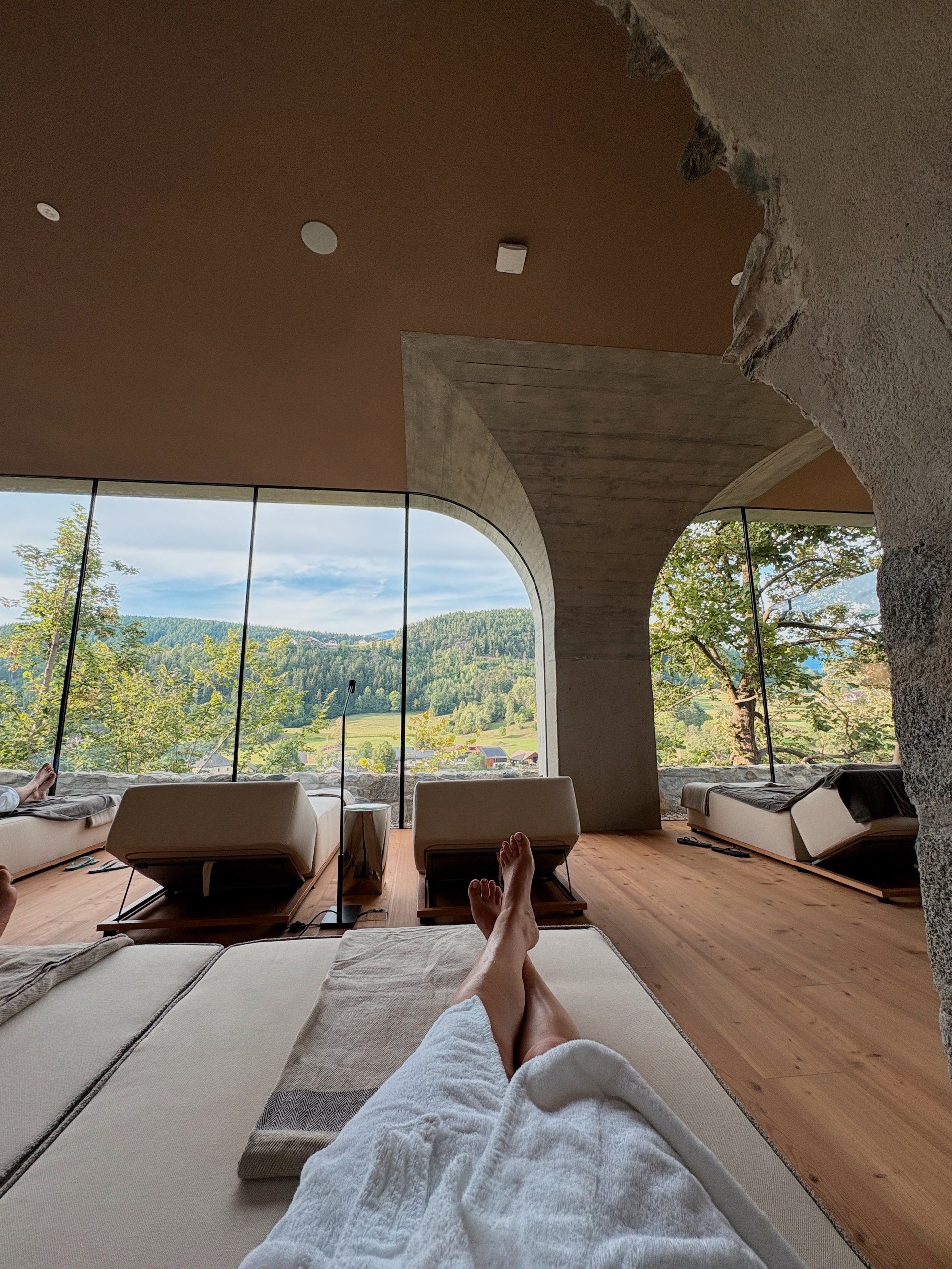 Guest in a white robe relaxing on a spa daybed facing floor-to-ceiling windows and a forested valley