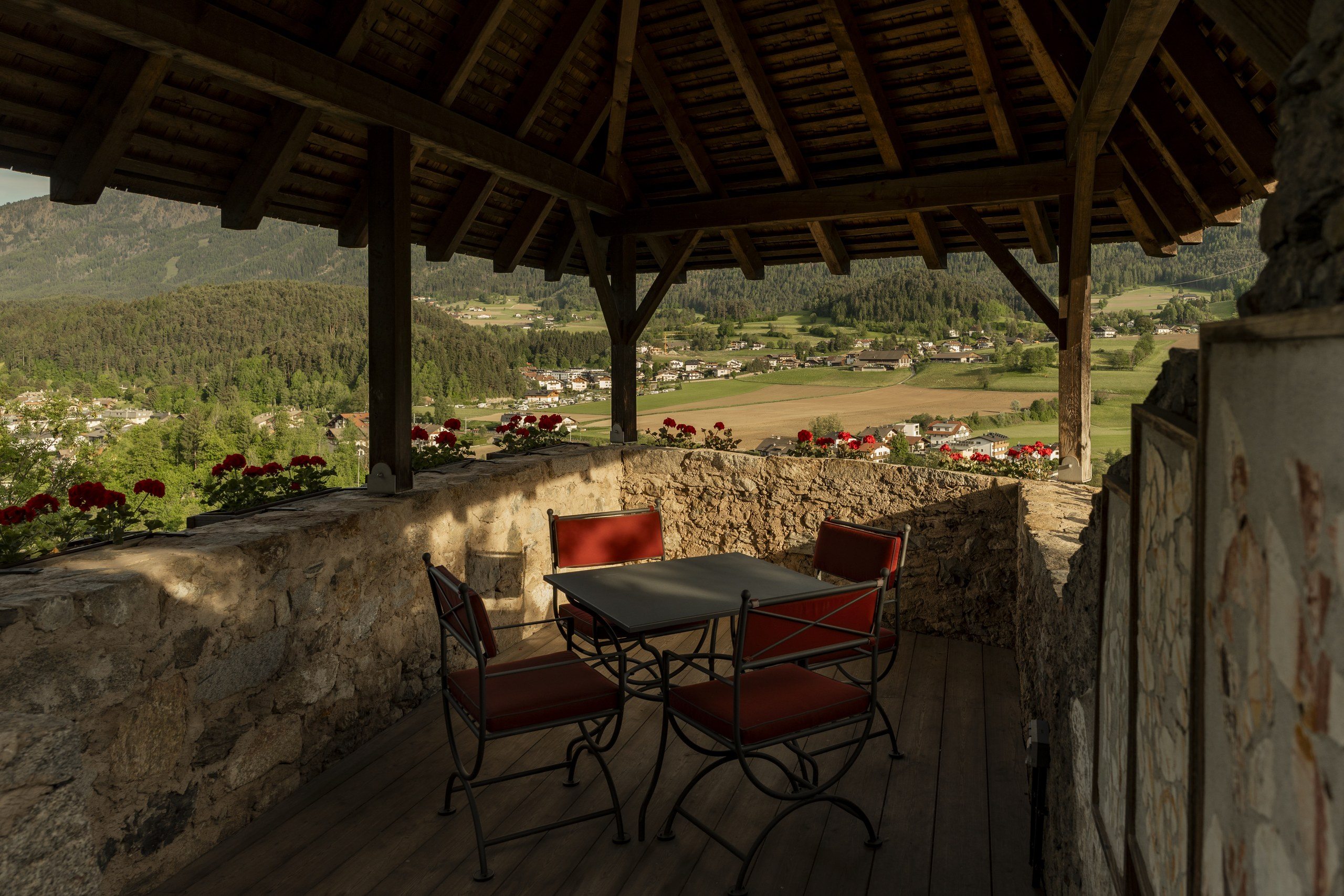 Covered stone terrace with a table for four and red geraniums overlooking a green Dolomites valley
